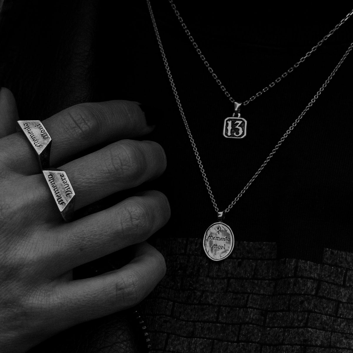 A close-up black and white photo of a person wearing layered silver necklaces and two silver Memento Mori signet-style rings on their fingers.