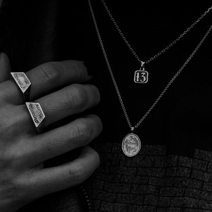 A close-up black and white photo of a person wearing layered silver necklaces and two silver Memento Mori signet-style rings on their fingers.