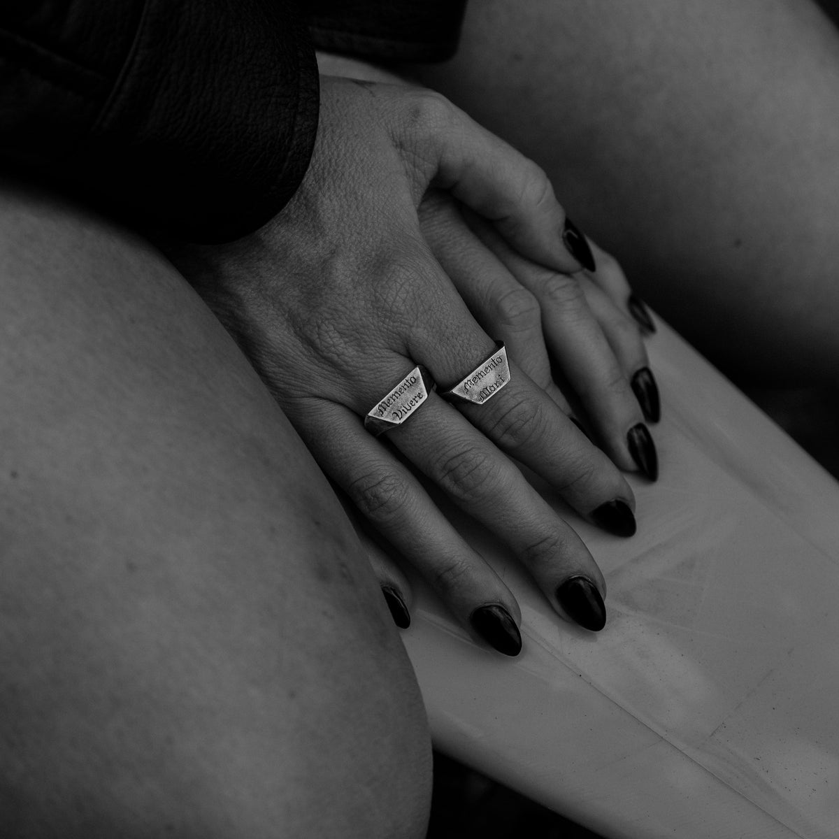 A close-up black and white photograph of a person's hands resting on their lap, wearing two memento sigent silver rings.