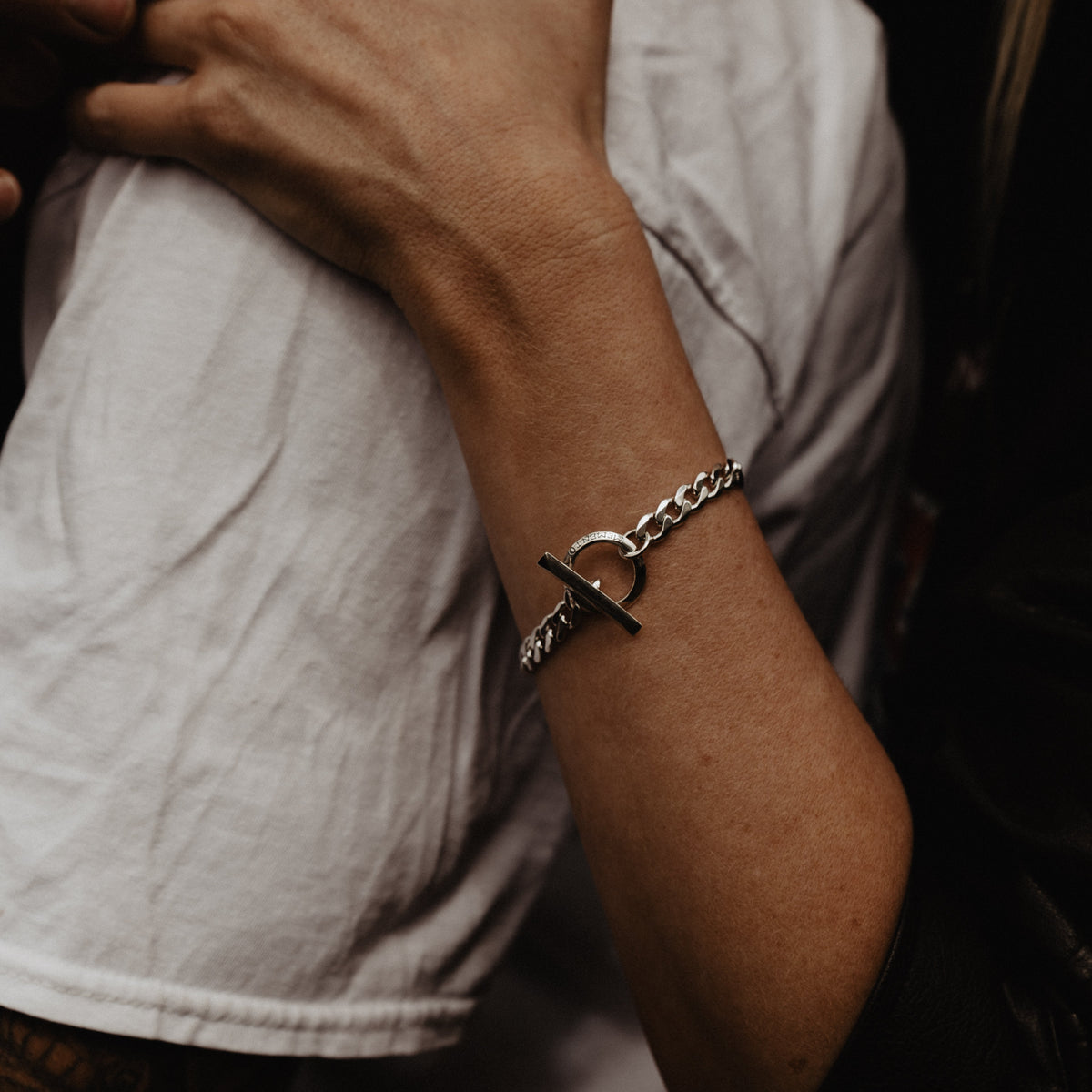 A close-up of a silver chain bracelet, shown against a dark background and white shirt.