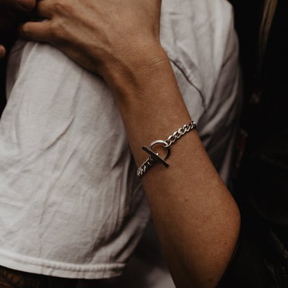 A close-up of a silver chain bracelet, shown against a dark background and white shirt.