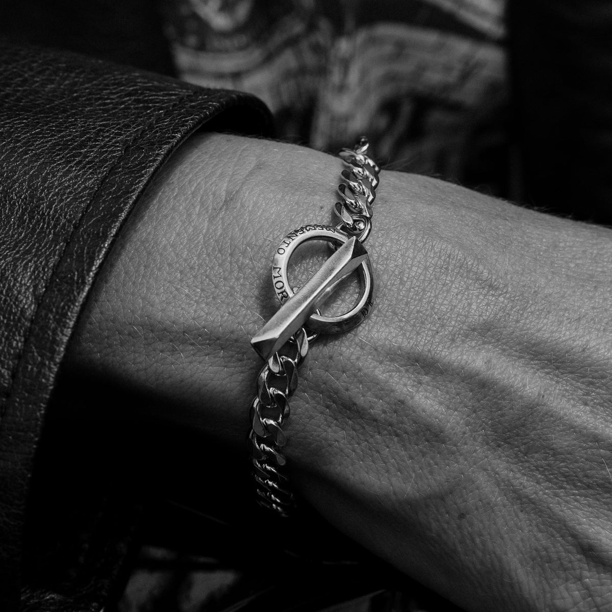 A close-up of a silver chain bracelet with a toggle clasp worn on a person's wrist, shown in black and white with a leather jacket cuff visible.